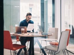 Lending alternativo: la nuova frontiera del credito per le PMI italiane. Uomo con occhiali lavora in un ufficio moderno leggendo un documento con grafici; sul tavolo laptop, tablet, caffè e cancelleria, in una sala riunioni con pareti in vetro.