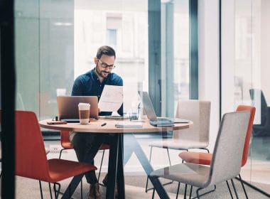 Lending alternativo: la nuova frontiera del credito per le PMI italiane. Uomo con occhiali lavora in un ufficio moderno leggendo un documento con grafici; sul tavolo laptop, tablet, caffè e cancelleria, in una sala riunioni con pareti in vetro.