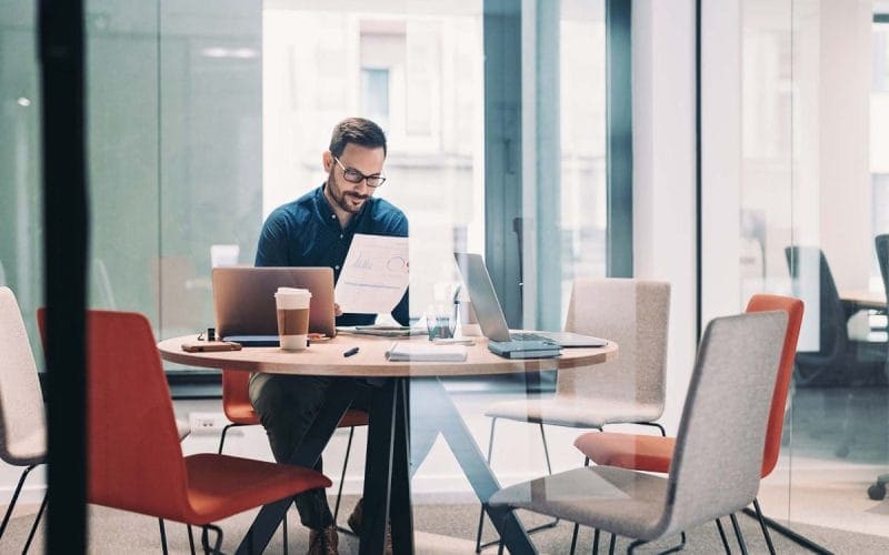 Lending alternativo: la nuova frontiera del credito per le PMI italiane. Uomo con occhiali lavora in un ufficio moderno leggendo un documento con grafici; sul tavolo laptop, tablet, caffè e cancelleria, in una sala riunioni con pareti in vetro.