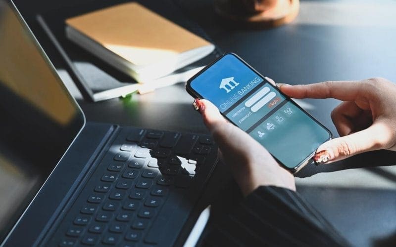 PMI e accesso al credito: perché il lending digitale sta cambiando le regole del gioco. Donna utilizza un’app di online banking su smartphone accanto a un laptop, simbolo della digitalizzazione finanziaria per PMI e liberi professionisti.