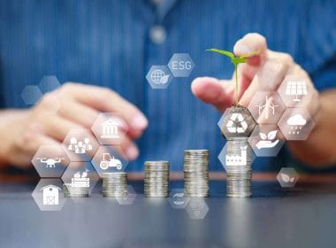 Mani posano una piantina su una pila di monete con icone ESG, a simboleggiare l’integrazione della sostenibilità nei criteri di accesso al credito aziendale.