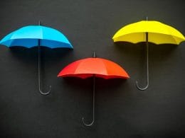 La differenza tra polizza vita e polizza infortuni: facciamo chiarezza. Tre ombrelli colorati — uno blu, uno rosso e uno giallo — su sfondo nero, simbolo visivo di protezione, copertura assicurativa e gestione dei rischi.