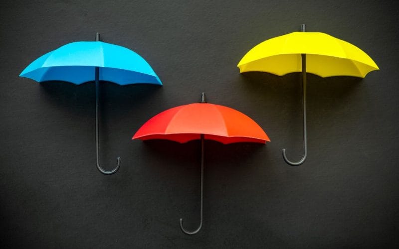 La differenza tra polizza vita e polizza infortuni: facciamo chiarezza. Tre ombrelli colorati — uno blu, uno rosso e uno giallo — su sfondo nero, simbolo visivo di protezione, copertura assicurativa e gestione dei rischi.