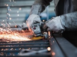 Lavori usuranti e pensione: quali possibilità di uscita anticipata per chi svolge attività particolarmente faticose? Operaio metalmeccanico con guanti protettivi utilizza una smerigliatrice, producendo scintille, simbolo di lavoro usurante e attività gravose ai fini pensionistici.