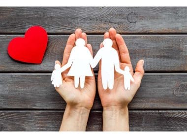 Le 5 polizze base che ogni famiglia dovrebbe valutare. Due mani che sorreggono una sagoma di famiglia con genitori e bambini, accanto a un cuore rosso su sfondo in legno: simbolo della protezione familiare e dell’assicurazione sulla vita.
