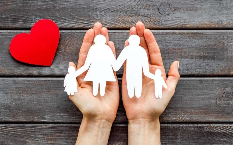 Le 5 polizze base che ogni famiglia dovrebbe valutare. Due mani che sorreggono una sagoma di famiglia con genitori e bambini, accanto a un cuore rosso su sfondo in legno: simbolo della protezione familiare e dell’assicurazione sulla vita.