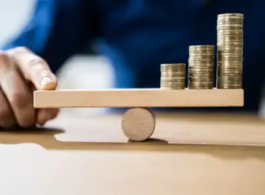 Perché assicurarsi è un investimento e non una spesa. Mano che bilancia una tavoletta con pile crescenti di monete su un fulcro centrale: concetto visivo di equilibrio finanziario e gestione ottimale delle risorse.