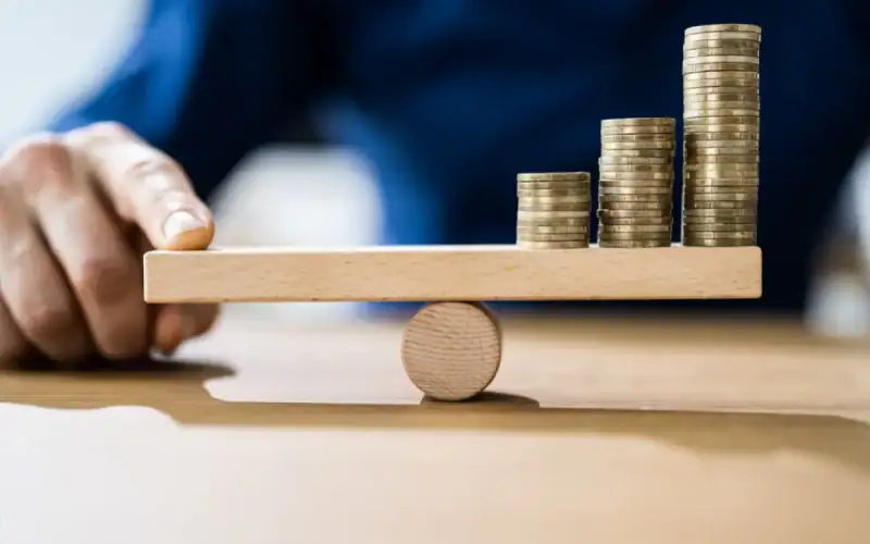 Perché assicurarsi è un investimento e non una spesa. Mano che bilancia una tavoletta con pile crescenti di monete su un fulcro centrale: concetto visivo di equilibrio finanziario e gestione ottimale delle risorse.