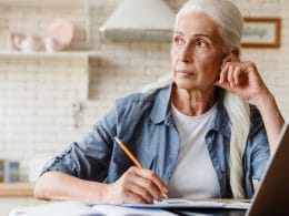 Pensione anticipata per lavoratori precoci: requisiti, categorie tutelate e regole di accesso. Donna matura con capelli bianchi seduta al tavolo, riflette mentre prende appunti con un laptop aperto davanti, simbolo di pianificazione della pensione anticipata per lavoratori precoci.