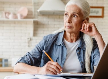 Pensione anticipata per lavoratori precoci: requisiti, categorie tutelate e regole di accesso. Donna matura con capelli bianchi seduta al tavolo, riflette mentre prende appunti con un laptop aperto davanti, simbolo di pianificazione della pensione anticipata per lavoratori precoci.