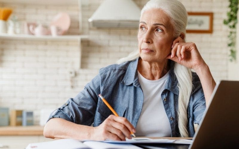 Pensione anticipata per lavoratori precoci: requisiti, categorie tutelate e regole di accesso. Donna matura con capelli bianchi seduta al tavolo, riflette mentre prende appunti con un laptop aperto davanti, simbolo di pianificazione della pensione anticipata per lavoratori precoci.