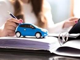 L’assicurazione auto in Italia: dalle origini alla rivoluzione digitale. Modellino di auto blu su un fascicolo di documenti, con una donna che firma o consulta un contratto, simbolo di leasing, finanziamenti auto o noleggio a lungo termine.