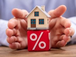 Bonus ristrutturazioni. Modellino di casa su un cubo rosso con simbolo di percentuale, circondato da mani protettive: rappresentazione di mutui, tassi d’interesse e sicurezza finanziaria legata alla casa.