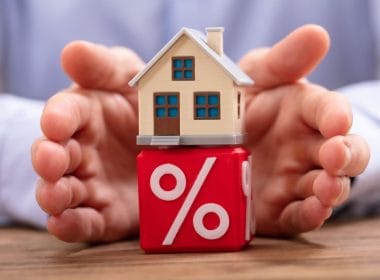 Bonus ristrutturazioni. Modellino di casa su un cubo rosso con simbolo di percentuale, circondato da mani protettive: rappresentazione di mutui, tassi d’interesse e sicurezza finanziaria legata alla casa.