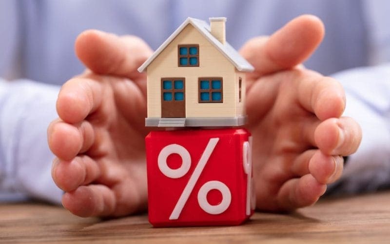 Bonus ristrutturazioni. Modellino di casa su un cubo rosso con simbolo di percentuale, circondato da mani protettive: rappresentazione di mutui, tassi d’interesse e sicurezza finanziaria legata alla casa.