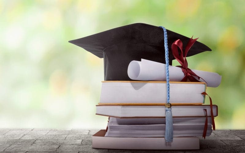 Riscatto della laurea: il riscatto ordinario conviene davvero? Pile di libri con tocco accademico e diploma arrotolato: simbolo del riscatto della laurea per ottenere anni contributivi validi per la pensione.