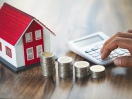 Aggiornamento della rendita catastale. Mano che utilizza una calcolatrice accanto a pile di monete, un modellino di casa e sfondo in legno, rappresentazione di calcolo mutuo, investimento e valore immobiliare.