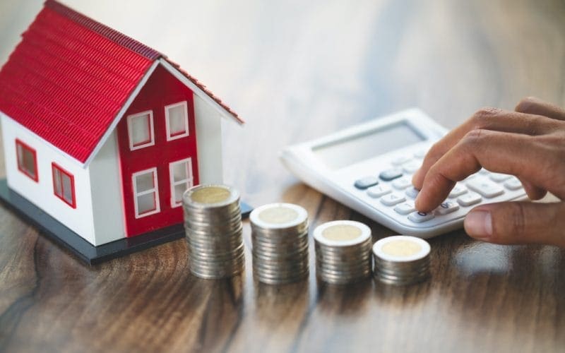 Aggiornamento della rendita catastale. Mano che utilizza una calcolatrice accanto a pile di monete, un modellino di casa e sfondo in legno, rappresentazione di calcolo mutuo, investimento e valore immobiliare.