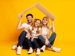 Cosa copre (davvero) una polizza sulla casa? Famiglia felice composta da padre, madre e figlia seduti su sfondo giallo con un tetto stilizzato sopra la testa, simbolo di protezione assicurativa della casa.