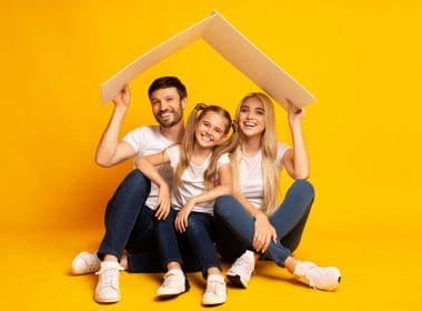 Cosa copre (davvero) una polizza sulla casa? Famiglia felice composta da padre, madre e figlia seduti su sfondo giallo con un tetto stilizzato sopra la testa, simbolo di protezione assicurativa della casa.