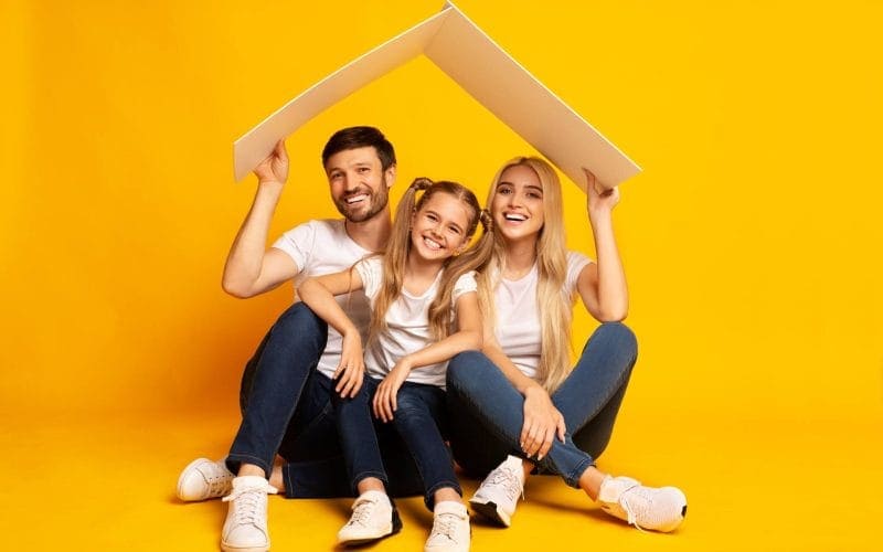 Cosa copre (davvero) una polizza sulla casa? Famiglia felice composta da padre, madre e figlia seduti su sfondo giallo con un tetto stilizzato sopra la testa, simbolo di protezione assicurativa della casa.