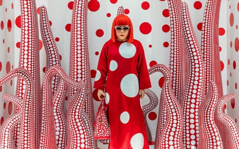 Mercato dell'arte. Donna con parrucca rossa in ambiente immersivo optical rosso e bianco, stile Yayoi Kusama, simbolo dell’arte contemporanea femminile.