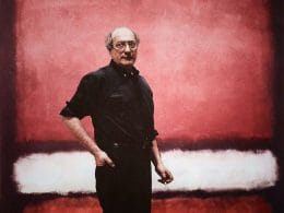 Arte come bene rifugio. Mark Rothko. Ritratto di Mark Rothko in piedi davanti a un suo dipinto astratto con campi di colore rosso, bianco e nero; artista in abiti scuri con espressione seria.