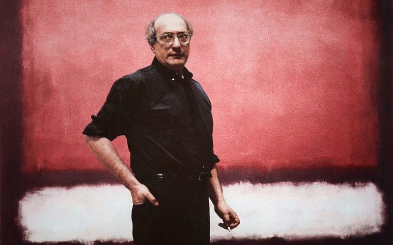 Arte come bene rifugio. Mark Rothko. Ritratto di Mark Rothko in piedi davanti a un suo dipinto astratto con campi di colore rosso, bianco e nero; artista in abiti scuri con espressione seria.