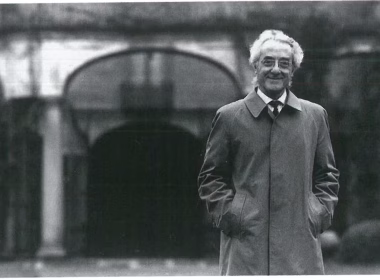 Grandi collezionisti italiani: Giuseppe Panza di Bium con cappotto elegante sorride davanti a un edificio con archi in stile classico; immagine in bianco e nero scattata all’esterno.