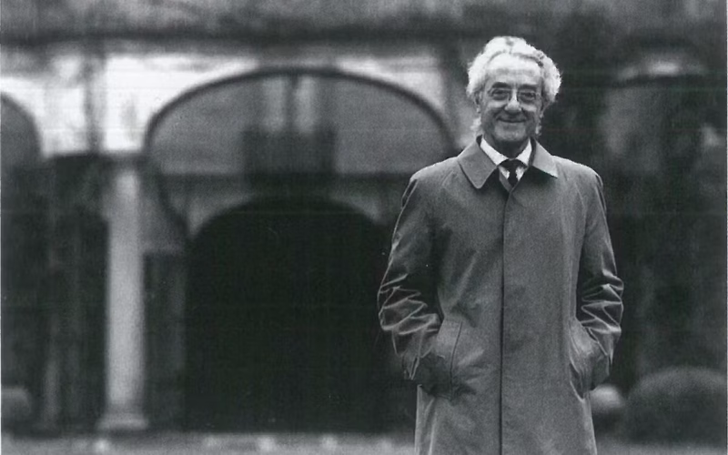 Grandi collezionisti italiani: Giuseppe Panza di Bium con cappotto elegante sorride davanti a un edificio con archi in stile classico; immagine in bianco e nero scattata all’esterno.