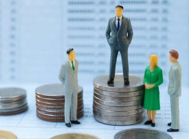 Crisi di liquidità per le PMI? Quattro miniature di persone in abiti da ufficio attorno a pile di monete, con una figura al centro posizionata su una pila più alta, simbolo di disparità economica tra PMI.