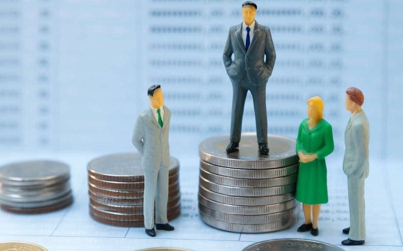 Crisi di liquidità per le PMI? Quattro miniature di persone in abiti da ufficio attorno a pile di monete, con una figura al centro posizionata su una pila più alta, simbolo di disparità economica tra PMI.