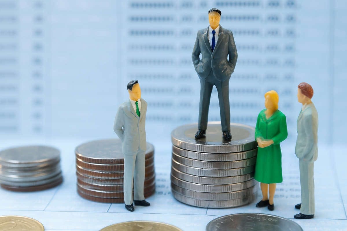 Crisi di liquidità per le PMI? Quattro miniature di persone in abiti da ufficio attorno a pile di monete, con una figura al centro posizionata su una pila più alta, simbolo di disparità economica tra PMI.