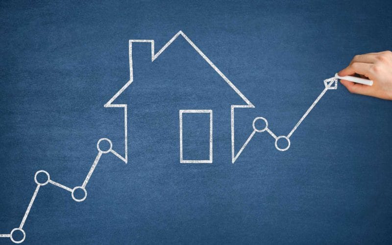 Crowdfunding immobiliare. Grafico a linea crescente con cerchi di raccordo e freccia finale, disegnato con gesso bianco su una lavagna blu, che attraversa una sagoma stilizzata di una casa.