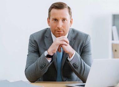Uomo in abito grigio, seduto a una scrivania moderna con un laptop, che guarda in camera con espressione determinata — rappresentazione simbolica del consulente del futuro.