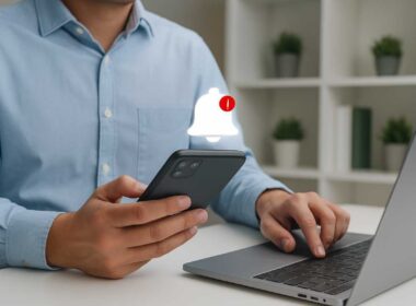Truffe Instagram e WhatsApp. Uomo che controlla notifiche sullo smartphone davanti a un laptop, simbolo di clone Instagram e possibili truffe online.
