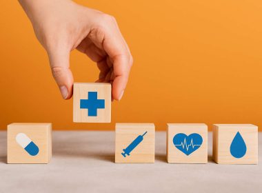 Blocchi di legno con simboli sanitari e mano femminile che solleva un cubo con croce blu, concetto di esigenze mediche insoddisfatte e difficoltà di accesso alle cure.