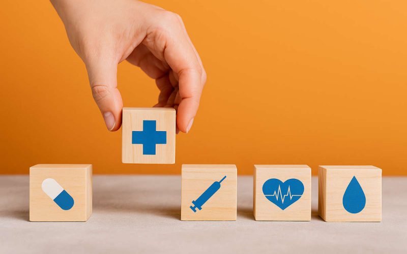 Blocchi di legno con simboli sanitari e mano femminile che solleva un cubo con croce blu, concetto di esigenze mediche insoddisfatte e difficoltà di accesso alle cure.