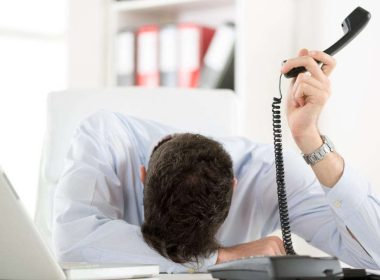 Uomo stanco e frustrato dalle troppe chiamate di telemarketing, con la testa sulla scrivania e il telefono in mano