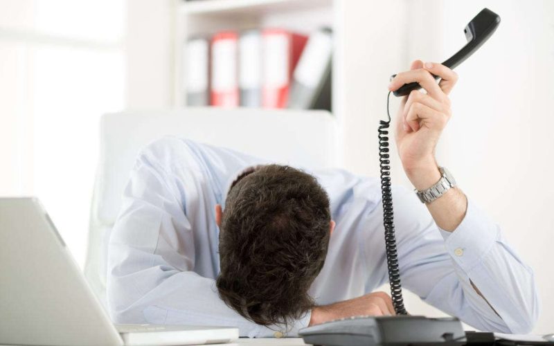Uomo stanco e frustrato dalle troppe chiamate di telemarketing, con la testa sulla scrivania e il telefono in mano