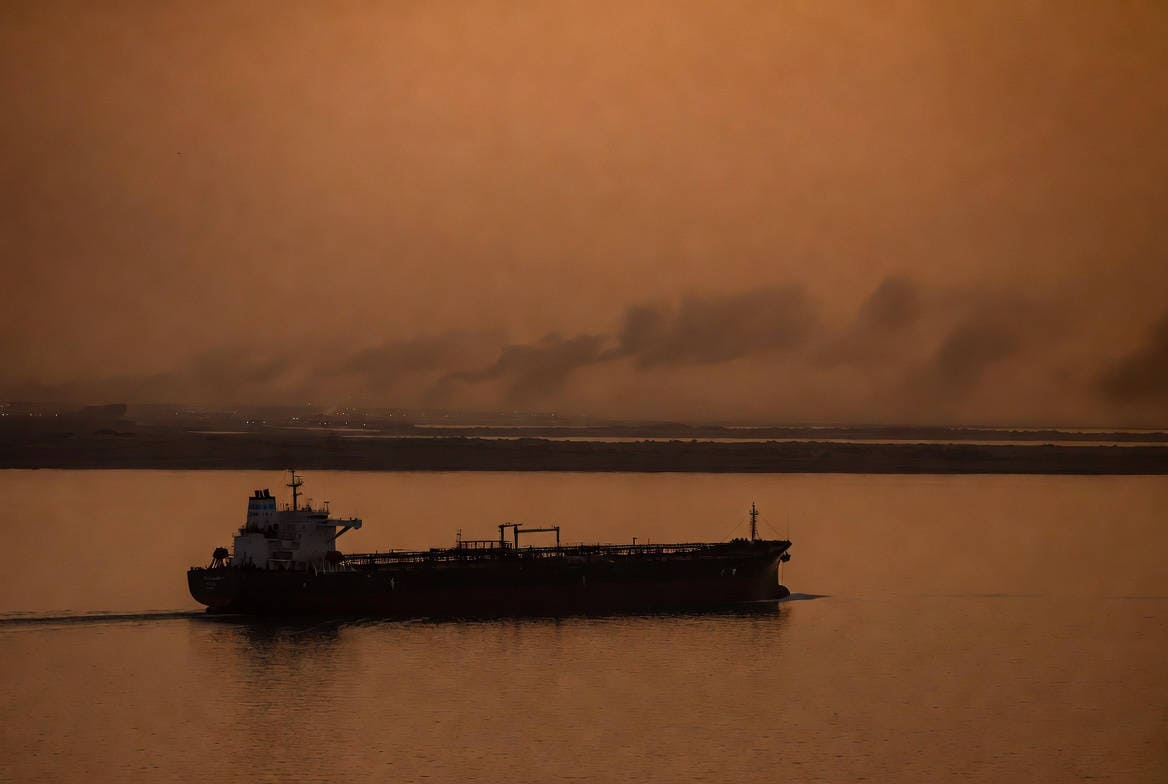 Crisi energetica e recessione 2026: petroliera ferma nello Stretto di Hormuz al crepuscolo con foschia ambrata.