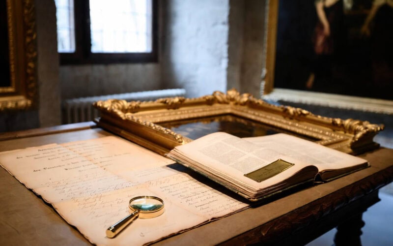Due diligence etica: come evitare riciclaggio, opere rubate e provenienza tossica. Verifica provenienza opere d'arte: documento manoscritto d'epoca, lente d'ingrandimento e catalogo su tavolo con cornice dorata