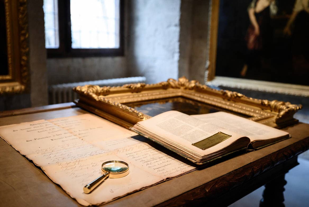 Verifica provenienza opere d'arte: documento manoscritto d'epoca, lente d'ingrandimento e catalogo su tavolo con cornice dorata