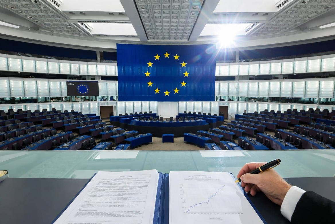 Savings and Investments Union: emiciclo del Parlamento europeo vuoto con bandiera UE e documenti finanziari in primo piano