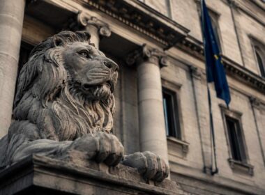 La banca presta ancora. Ma non a te.. accesso al credito per le PMI, statua di leone davanti a un edificio istituzionale con colonne e bandiera europea